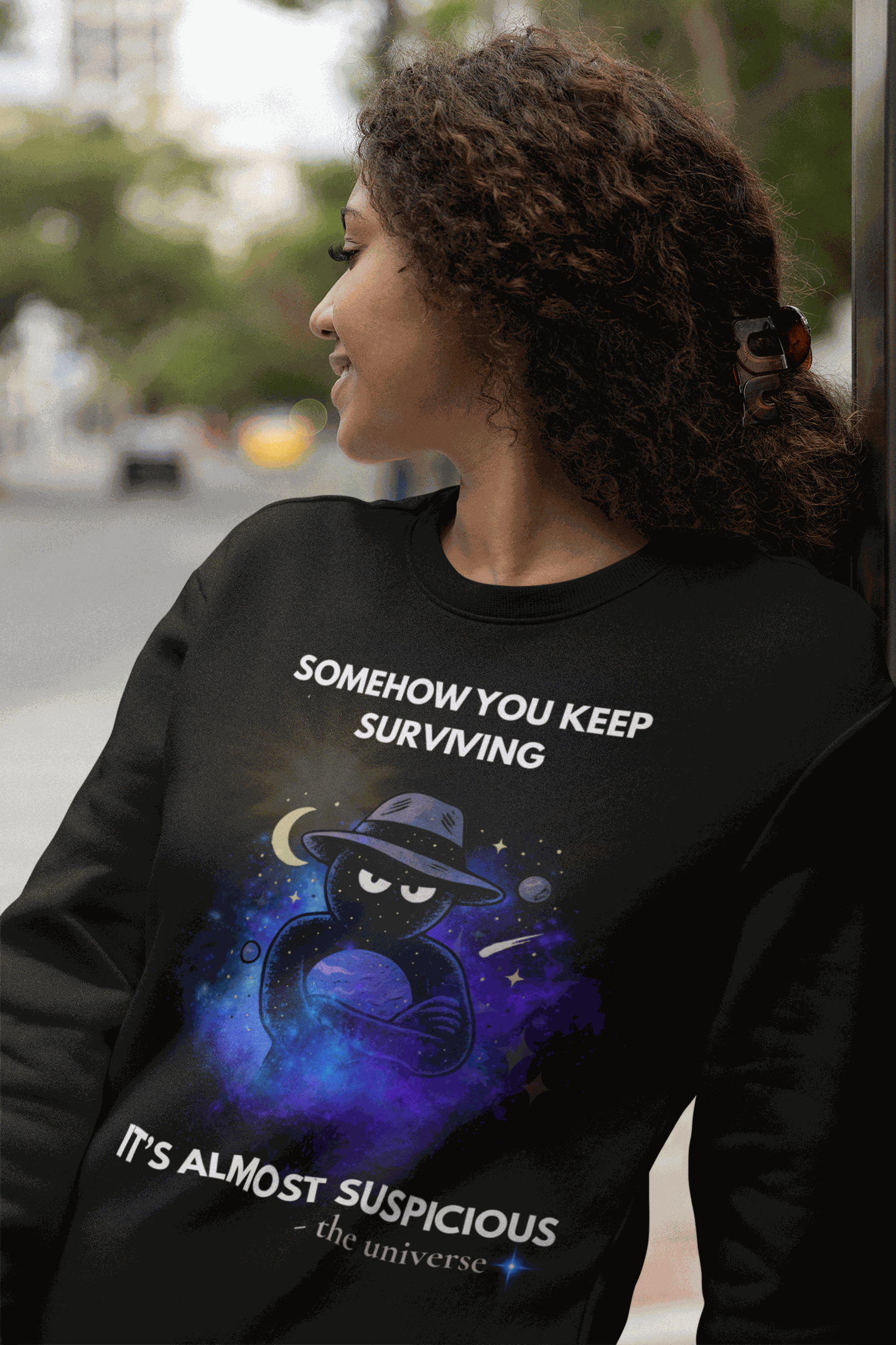 A black sweatshirt with a cosmic graphic and the text "Somehow you keep surviving, it's almost suspicious".