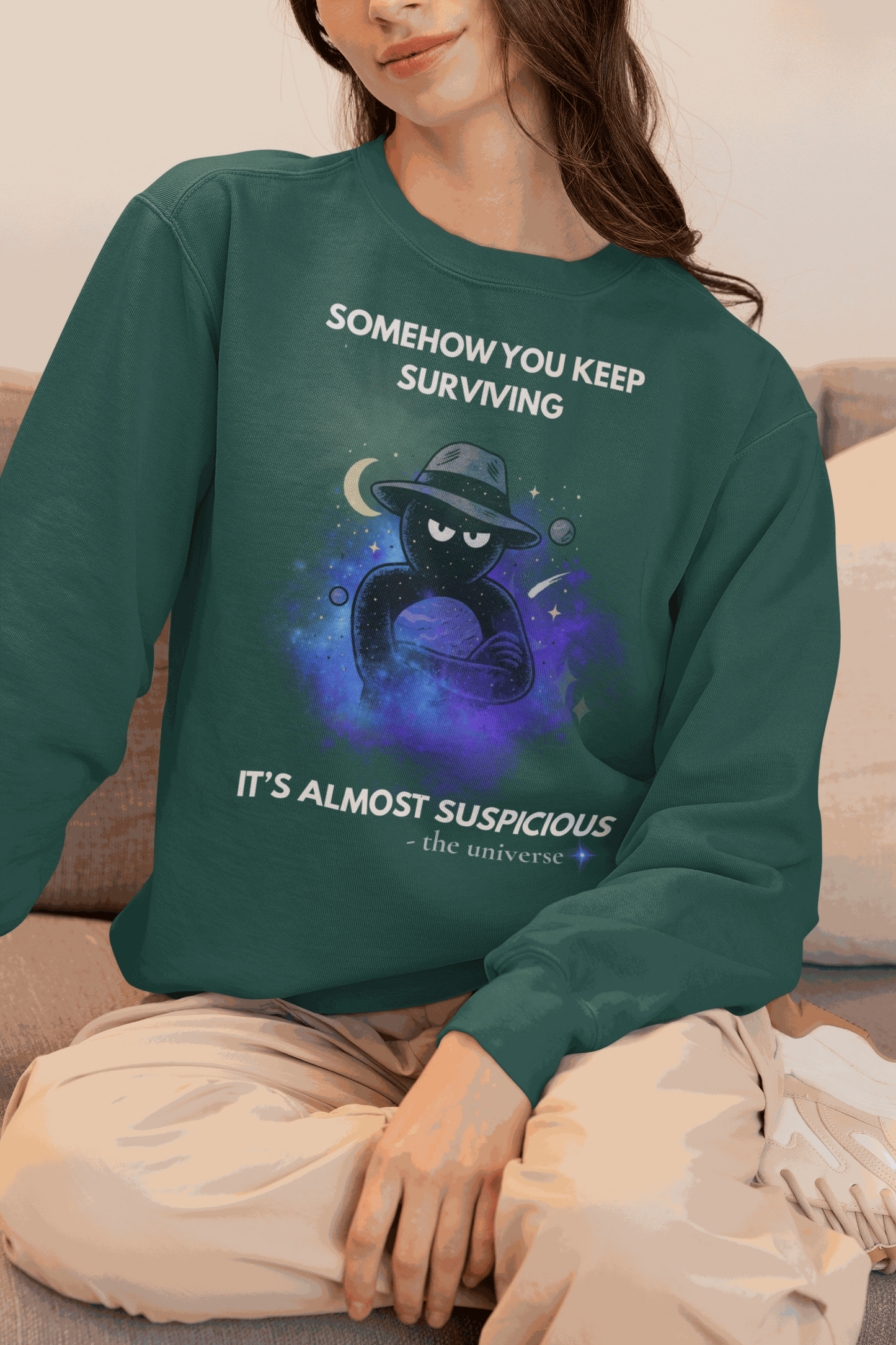 A dark green sweatshirt with a cosmic graphic and the text "Somehow you keep surviving it's almost suspicious".