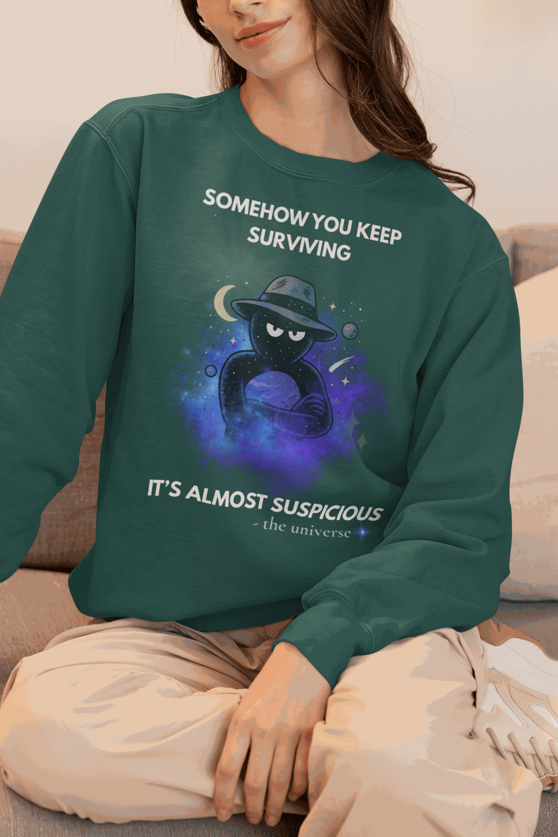 A dark green sweatshirt with a cosmic graphic and the text "Somehow you keep surviving it's almost suspicious".
