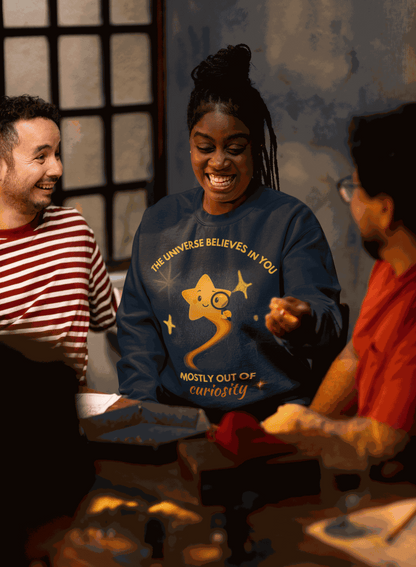 A dark blue sweatshirt with a graphic of a star looking through a magnifying glass and the text "The Universe Believes in You Mostly Out of Curiosity".