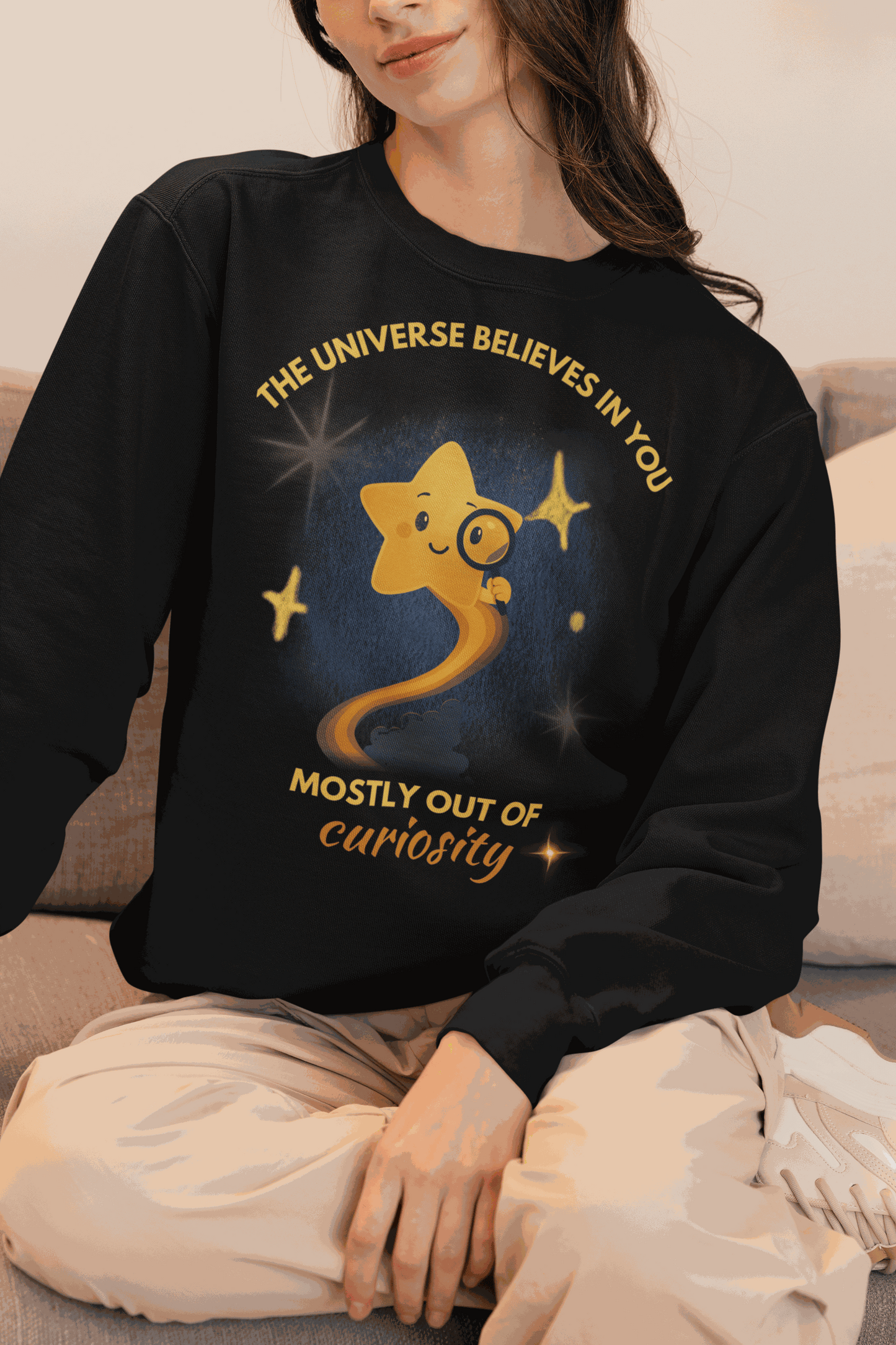 A black sweatshirt with a graphic of a star looking through a magnifying glass and text that reads "The Universe Believes in You Mostly Out of Curiosity".