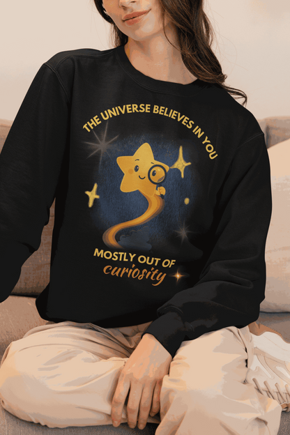 A black sweatshirt with a graphic of a star looking through a magnifying glass and text that reads "The Universe Believes in You Mostly Out of Curiosity".