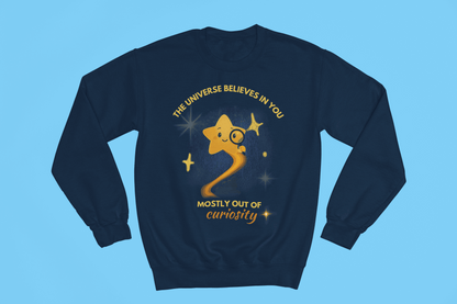 A navy sweatshirt with a graphic of a star looking through a magnifying glass and text that reads "The Universe Believes in You, Mostly Out of Curiosity".