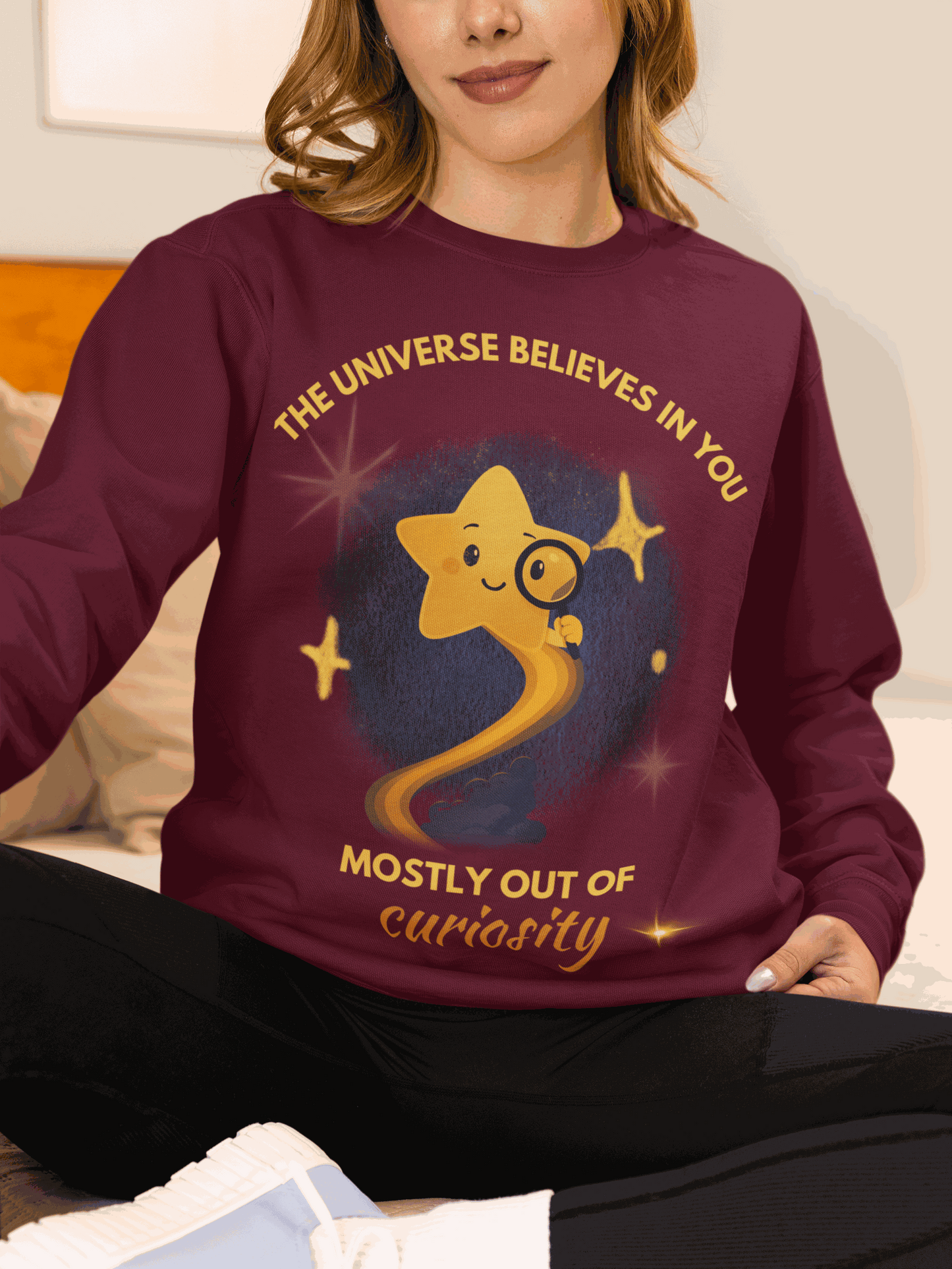A maroon sweatshirt with a graphic of a star and the text "The Universe Believes in You Mostly Out of Curiosity".