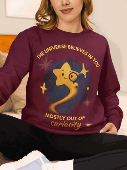 A maroon sweatshirt with a graphic of a star and the text "The Universe Believes in You Mostly Out of Curiosity".